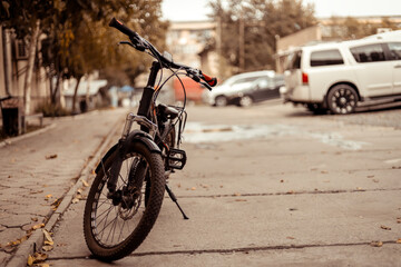 Black bike parked on a city sidewalk with its front wheel facing the viewer. The bicycle is leaning on its kickstand. The sidewalk has a few fallen leaves on it.