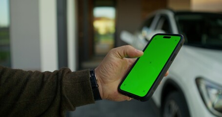 A man uses a mobile phone with green screen standing near an electric car. Close-up of his hands.