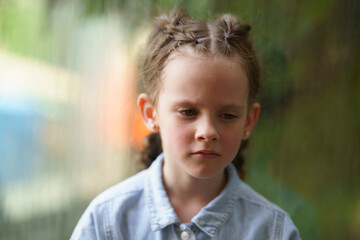 Little sad girl with two big pigtails near the glass. Concept of sad childhood