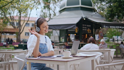 Cheerful girl greeting video call laptop at street cafe in headphones closeup.