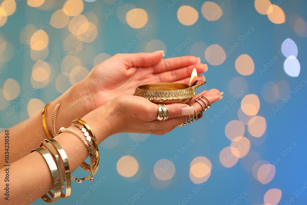 Sticker Diwali celebration. Woman holding lit diya lamp on light blue background with blurred lights, closeup