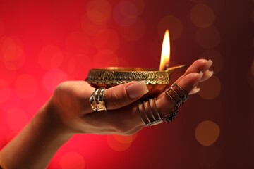 Diwali celebration. Woman holding lit diya lamp on red background with blurred lights, closeup