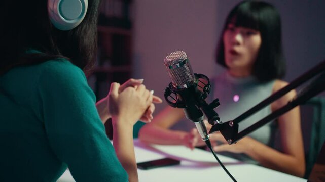 Radio host wearing headphones gestures while conducting an engaging interview with a guest in a professional recording studio, surrounded by audio equipment and microphones
