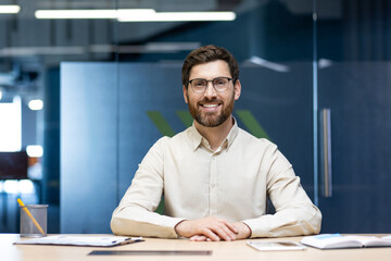 Portrait of a smiling confident young businessman in a light shirt and glasses sitting at a desk in...
