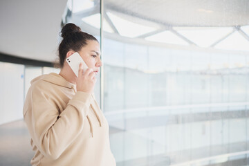 Emotions and facial expressions: serious face of a young woman with a bun in her hair listening to someone on a mobile phone next to a hospital window.