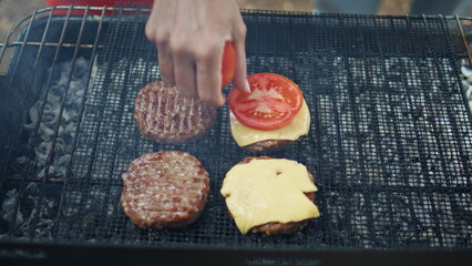 Closeup hand adding tomato cheese to burger patties on grill at park picnic. 