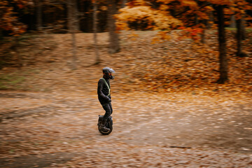 A man wearing protective gear rides an electric unicycle across a forest path covered in autumn leaves, with blurred motion effect. © Raivo