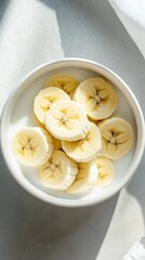A white bowl contains freshly sliced bananas with a bright, appealing appearance. The scene is illuminated by soft, natural light, creating a refreshing feel