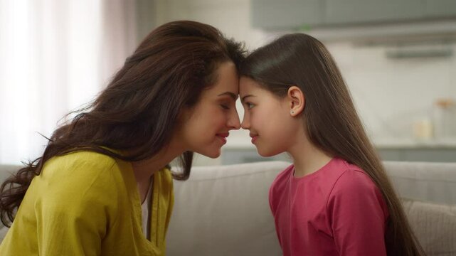 Cute love moment family bonding Caucasian woman female mother touching forehead cuddling with little girl preschool child daughter kid hugging on sofa embracing hug motherhood at home cozy apartment