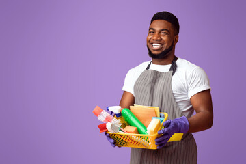 Professional Cleaner. Positive black cleaning service worker holding basket with household supplies and smiling at camera, pink background