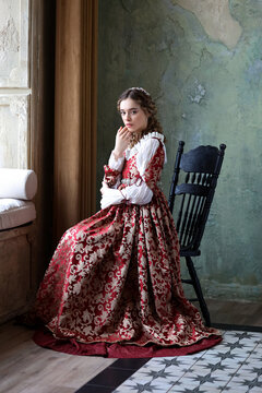 Young woman in historic renaissance dress with curled hairstyle sitting in old room with antique interior. Historical portrait of medieval maiden.