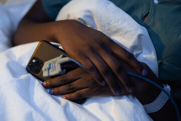 Fototapeta premium Close up shot on African patient's hands with phone in hands and oximeter in hospital ward.