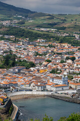 Aerial view of historic town on Terceira Island, Portugal. The image captures the town's red-tiled roofs, coastal area, and lush green hills in the background.