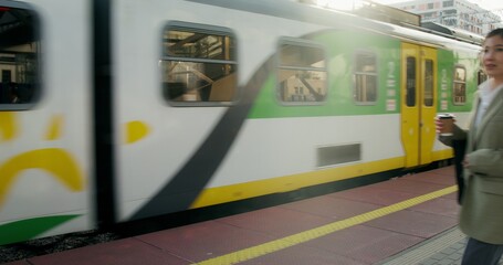 A woman in formal clothes, with headphones on her head and a cup of coffee in her hands is walking along the railway platform. The train passes by her