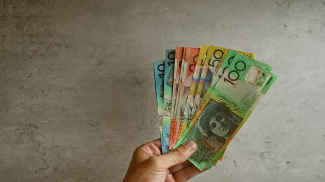 Hand holding a fan of australian dollars against a concrete backdrop, showcasing vibrant, colorful banknotes of various denominations.