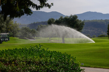 A serene golf course landscape with sprinklers at work under a clear sky in the early afternoon, enhancing the lush greenery and peaceful atmosphere