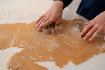 A handsome dark-haired boy with beautiful eyes and a smile in New Year's pajamas, rolls out gingerbread dough in the kitchen with a rolling pin, a Christmas tree stands in the background and a garland