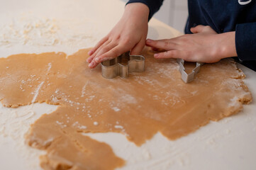A handsome dark-haired boy with beautiful eyes and a smile in New Year's pajamas, rolls out gingerbread dough in the kitchen with a rolling pin, a Christmas tree stands in the background and a garland