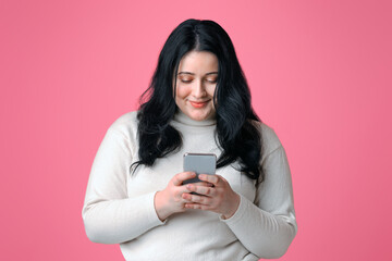 Smiling plump woman holding smartphone in hands, texting, reading message, or using new...