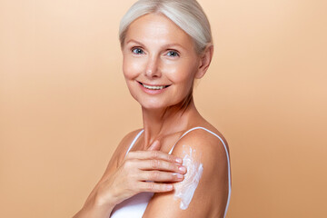 Beautiful mature woman with gray hair applying skincare lotion to her shoulder, highlighting self-care and age-positive beauty on a beige background.
