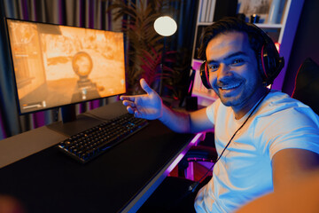 Smart gaming streamer looking at camera making self photo shooting with showing against with battle game on pc screen, using headset and mic talking others at digital neon light studio room. Surmise.