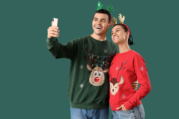 Young couple in Christmas sweaters and reindeer horns taking selfie on green background