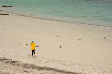 Teenager in yellow jacket walking on a white stone beach by turquoise ocean. Dog bay, county Galway, Ireland. Travel and tourism. Stunning Irish landscape scene. Enjoy nature theme.