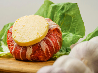 A beef burger wrapped in bacon or rasher and a slice of cheese on top of it and garlic cloves. The burger is surrounded by lettuce. The burger is on a wooden cutting board. High quality meat product