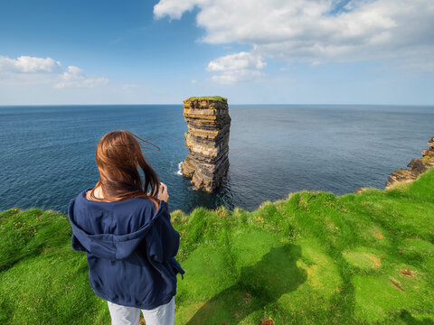 Teenager girl enjoys and films Dun Briste sea stuck, Downpatrick head, Ireland. Popular tourist attraction with stunning nature scenery. Warm sunny day. Travel and sightseeing Irish landmarks.