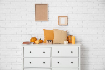 Chest of drawers with cushions, pumpkins and candle near white brick wall