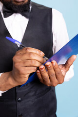 African american hotel porter making a list with room reservations to keep track of bookings and clients, blue background. Receptionist with suit and tie takes notes on papers. Close up.