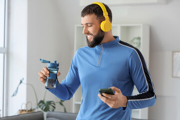 Sporty young man with mobile phone, headphones and water bottle at home