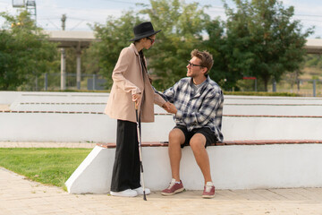 A man gently takes the hand of a blind woman while she stands beside him on a park bench. They share a moment of trust and compassion in a serene outdoor setting