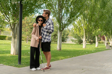 A caring man hugs a blind woman as they enjoy a leisurely walk in a vibrant park filled with greenery and sunshine, connecting through their shared moment