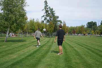 Two men are outdoors in a park, practicing their soccer juggling skills. One man performs ball tricks while the other watches, enjoying the sunny day among the trees