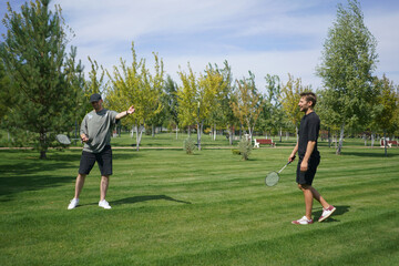 Fototapeta premium In a lush park, two friends play badminton on a beautiful sunny day. The vibrant greenery and clear sky create a perfect backdrop for their friendly match