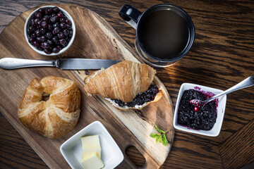 A plate of croissants and a cup of coffee sit on a wooden cutting board. The croissants are arranged in a way that they look like they are ready to be eaten with fresh wild berry jam.