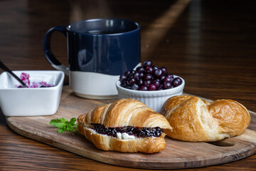A plate of croissants with huckleberry jam and a cup of coffee sit on a wooden table. The croissants are arranged on a wooden tray, and the coffee is in a blue mug. Concept of comfort and relaxation