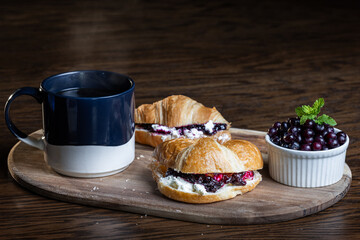 A plate of croissants with huckleberry jam and a cup of coffee sit on a wooden table. The croissants are arranged on a wooden tray, and the coffee is in a blue mug. Concept of comfort and relaxation