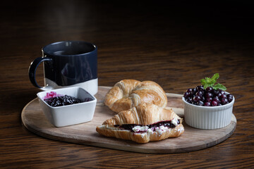 A plate of croissants with huckleberry jam and a cup of coffee sit on a wooden table. The croissants are arranged on a wooden tray, and the coffee is in a blue mug. Concept of comfort and relaxation
