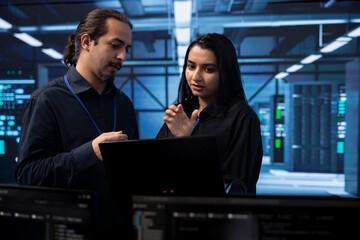 Technicians in data center using notebook to check for server performance issues, brainstorming solutions. Coworkers in server hub workspace fixing insufficient memory errors slowing down hardware