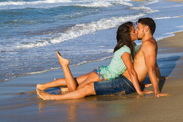 Happy young couple in bikini and shorts enjoying summer at the beach at dusk, breathing fresh air, kissing and teasing one another