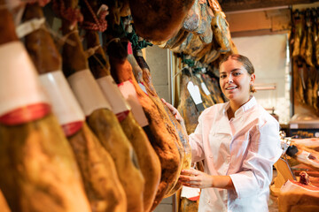 Portrait of positive female butcher on background of rack with hanging various jamon in store