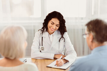 Consultation Concept. Happy latina doctor taking notes about symptoms, filling out medical prescription to senior couple