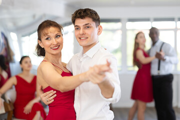 Happy smiling woman dressed in red dress enjoying waltz with male partner during group party in...
