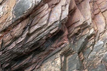 Close-up view of stratified rock formations, featuring layers of various shades of brown, reddish, and grey tones. The rugged texture and visible cracks highlight the natural geological processes