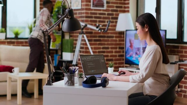 Software developer types programming code in a cozy home office setup. Asian woman coding for cloud computing on her laptop, enhances productivity for software development. Camera B.