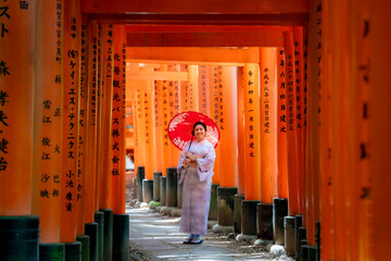 Japan, Kyoto - August 12, 2024: A young happy Latina woman in a pastel kimono, holding a red umbrella, smiles at Fushimi Inari-taisha shrine in Kyoto, Japan, surrounded by vibrant Torii gates, Japan