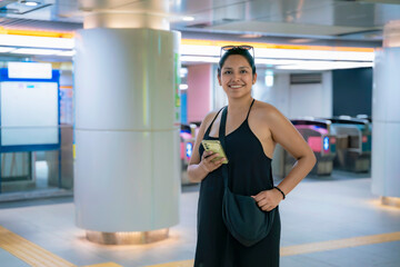 Smiling happy Latina woman in a black outfit smiles while holding her phone at Tokyo metro station, Japan