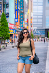 Latina happy woman smiling in sunglasses and a casual outfit surrounded by vibrant city life on busy Shinjuku street, Tokyo, Japan